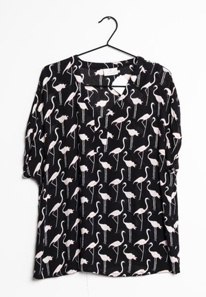 Blouse noire avec motif flamant rose clair suspendue à un cintre noir sur fond blanc.