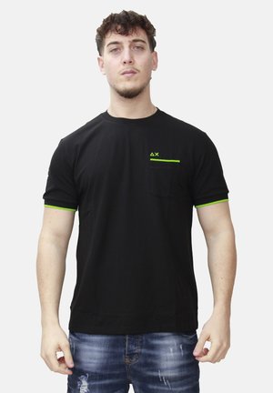 Giovane uomo che indossa una maglietta nera con rifiniture verde neon sulle maniche e tasca, abbinata a jeans blu strappati, in piedi davanti a uno sfondo bianco.