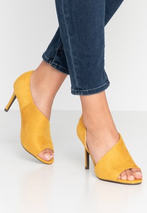Żółte zamszowe sandały na wysokim obcasie z otwartym przodem, charakteryzujące się krzyżującym się designem i eleganckim, spiczastym kształtem.