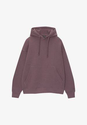 Person trägt einen lila Kapuzenpullover, weite, hellblaue, locker sitzende Jeans und graue Turnschuhe mit weißen Schnürsenkeln, steht mit den Händen in den Taschen.