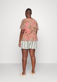 Person med kort hår iført en pink blomstrede bluse, mønstrede hvide og grå shorts, ogcremefarvede slip-on sko som står med ryggen til.