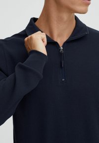 Pull-over zippé bleu marine en tissu texturé. Il possède un col et une fermeture éclair à l'avant avec tirette, mettant en avant un design structuré.