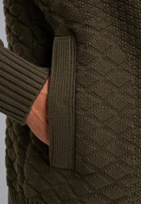 Maglione lavorato a maglia in un verde oliva scuro con un motivo a diamante tessuto, con polsini a coste e una tasca laterale visibile con una mano.