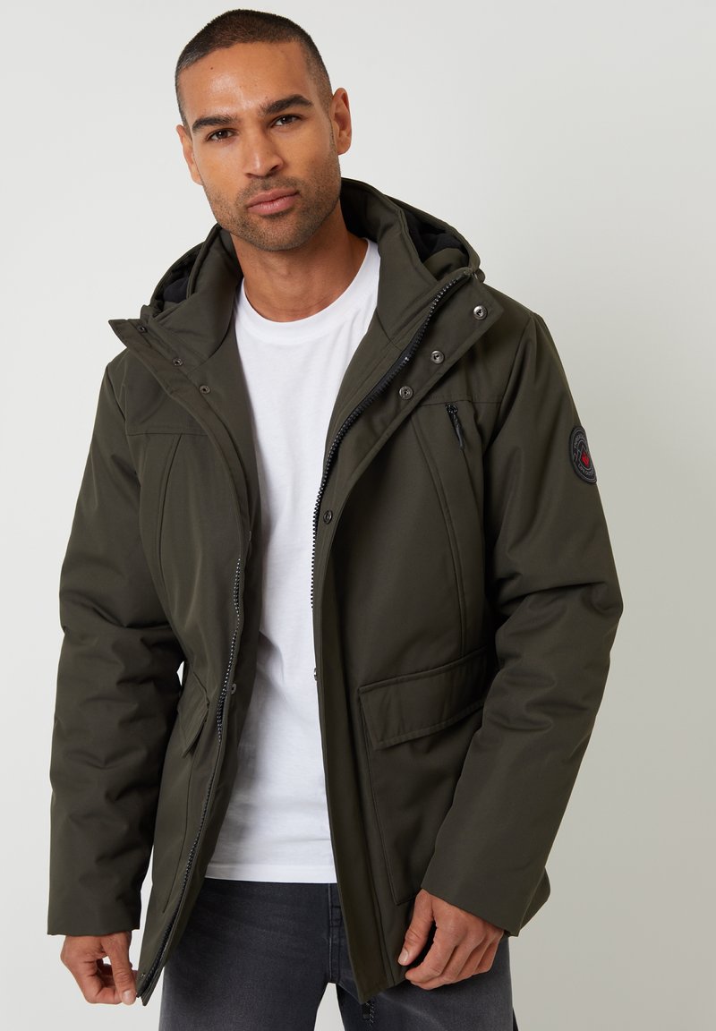 Dunkelgrüne isolierte Jacke mit Kapuze, Reißverschluss vorne und mehreren Taschen. Sie verfügt über einen strukturierten Stoff und ein Logopatch am Ärmel.