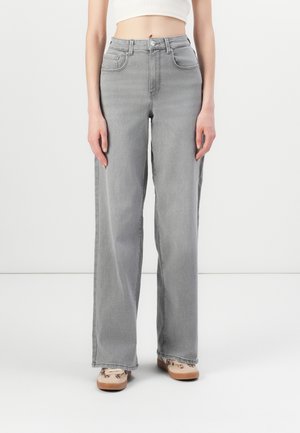 Woman wearing high-waist, wide-leg gray jeans and beige sneakers with abstract designs, standing against a white background.