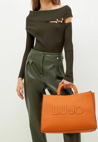 Orange tekstureret håndtaske med afrundede håndtag, med præget "LIU•JO" logo. Modellen har en ribbet mørkegrøn top og højtaljede grønne bukser på.