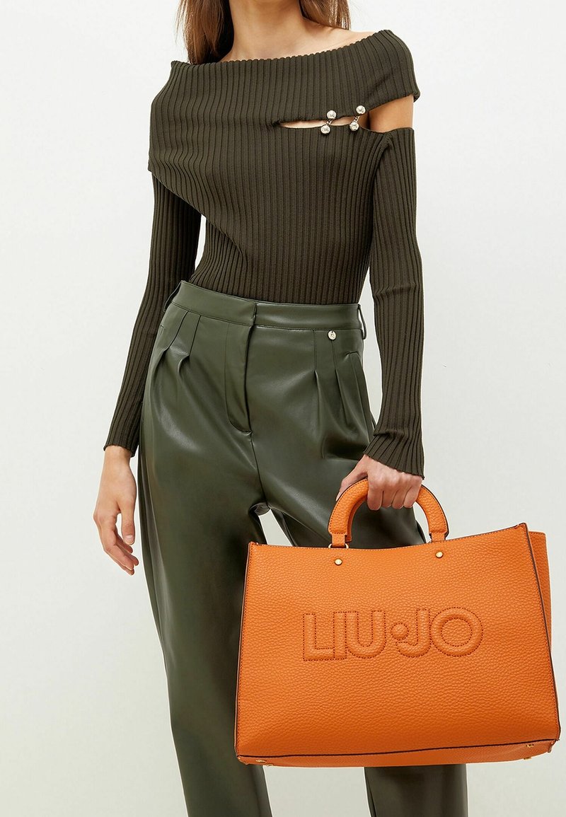 Orange tekstureret håndtaske med afrundede håndtag, med præget "LIU•JO" logo. Modellen har en ribbet mørkegrøn top og højtaljede grønne bukser på.