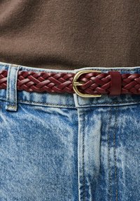 Nodrošināts sadruknāts bordo ādas jostiņs ar zelta metāla sprādzi, nēsāts virs gaiši zilo džinsām. Jostiņai redzams teksturēts raksts.