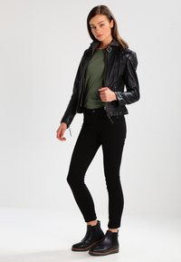 Black leather motorcycle jacket with textured shoulders, worn over a dark green T-shirt and black skinny jeans. Black ankle boots complete the look.