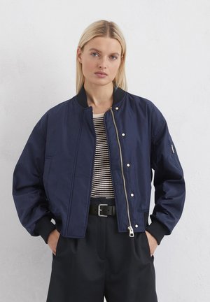 Navy bomber jacket with zip closure, ribbed cuffs, and side pockets. Worn over a striped top and wide black trousers. Smooth texture.