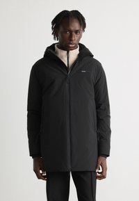 Doudoune noire avec capuche, dotée d'une fermeture éclair à l'avant, de poches latérales et d'un petit logo sur la poitrine. Portée par-dessus un haut en polaire beige.