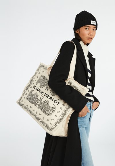 Gran bolsa tote de lona con un color crema y estampado de paisley negro, además de un logo. El modelo lleva puesta una chaqueta negra y un suéter a rayas.