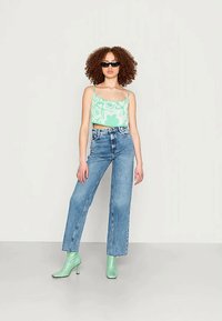 Top cropped floral verde, jeans de cintura alta azul-claro e botas altas verdes. Cabelo texturizado e óculos de sol escuros completam o visual.