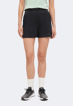Frau trägt schwarze, maßgeschneiderte Shorts, weiße Socken und graue Nike-Sneaker und steht vor einem einfarbigen hellen Hintergrund.