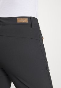 Zwarte rekbare broek heeft een gladde, matte textuur, een achterzak met rits en een bruine leren merklabel op de tailleband.