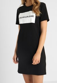 Černé tričko dress s krátkým rukávem s bílým obdélníkovým potiskem loga "CALVIN KLEIN JEANS" přes hruď. Měkká tkanina.