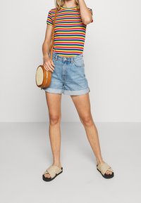 Camiseta a rayas multicolor, pantalones cortos de mezclilla con dobladillos doblados, bolso cruzado color marrón claro y sandalias beige con suelas negras.