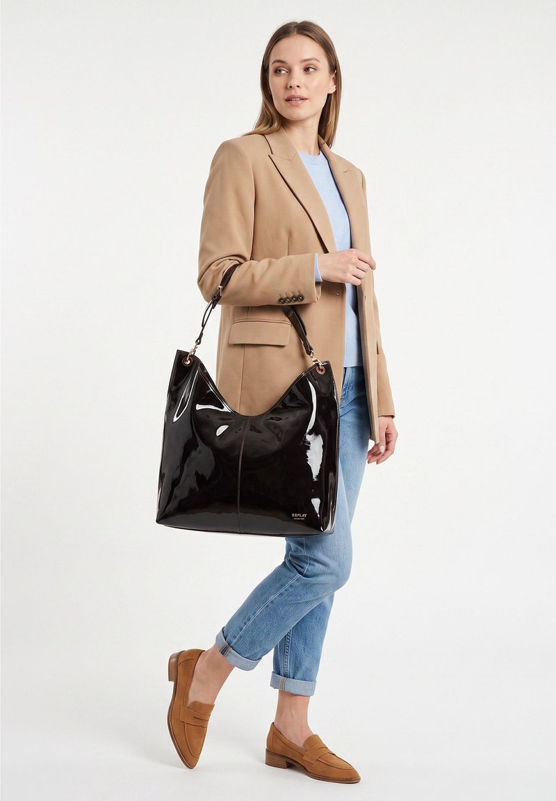 Femme portant un blazer beige, un jean bleu et des mocassins marron, tenant un grand sac à épaule noir en cuir verni, debout sur fond blanc.