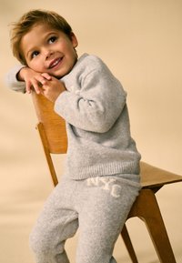 Maglione grigio in maglia e pantaloni coordinati con dettaglio "NYC", realizzati in materiale morbido. Bambino seduto su una sedia di legno con sfondo neutro.