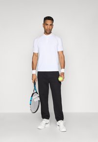 White short-sleeved athletic polo with a buttoned collar, paired with black textured track pants. Holds a tennis racket and ball.