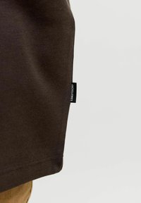 Gros plan sur un tissu marron foncé avec une petite étiquette noire portant la mention "JACK&JONES" sur la couture latérale sur un fond clair.