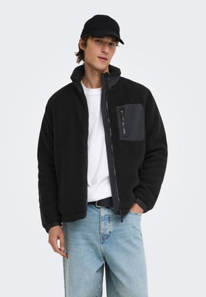 Jeune homme portant une casquette noire, une veste polaire noire zippée avec une poche poitrine, un t-shirt blanc et un jean bleu clair, debout devant un fond uni.