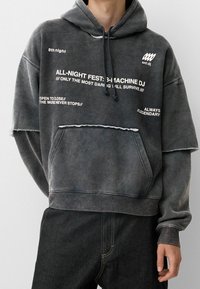 Sudadera gris de gran tamaño hecha de tela suave, con bolsillo frontal, bordes desgastados y gráficos de texto en blanco. Puños y dobladillo acanalados visibles.