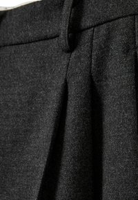 Gros plan d’un pantalon en tissu de laine gris foncé montrant la ceinture, les passants de ceinture et le détail plissé à l’avant.