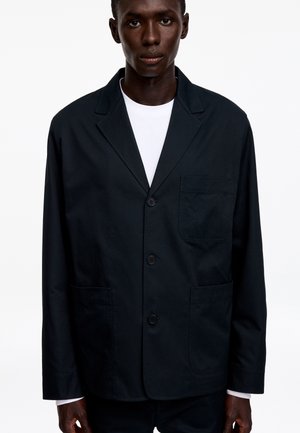 Homme portant un blazer noir boutonné avec trois poches devant sur une chemise blanche, sur un fond blanc uni.