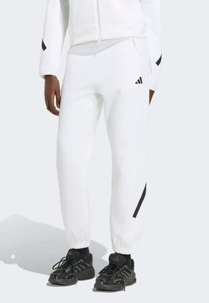 Osoba nosící bílé tepláky Adidas s černým logem, černé sportovní boty a bílou bundu na světlém pozadí.