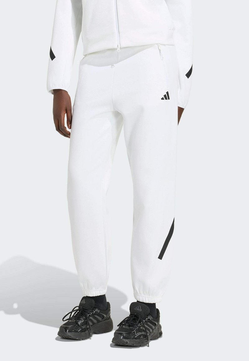Personne portant un pantalon de survêtement Adidas blanc avec logo noir, des chaussures de sport noires et une veste blanche sur un fond clair.
