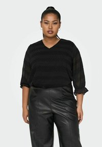 Blusa nera con scollatura a V e maniche tre quarti, caratterizzata da un motivo testurizzato. Abbinata a pantaloni in pelle nera a vita alta.