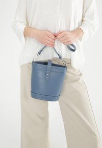Blauwe leren emmer tas met een gestructureerde afwerking, zilverkleurige hardware en een bovenhandvat. Draagt door een persoon in beige broek en een witte top.