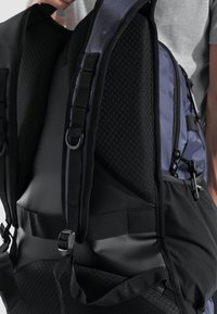 Sort og marineblå rygsæk med tekstureret mesh-bagside, justerbare stropper, lynlåse og webbing-detaljer. Holdbart materiale med polstrede sektioner.