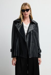 Donna con giacca oversize in ecopelle nera, blusa bianca, pantaloni larghi neri, occhiali cat-eye e grandi orecchini a cerchio, su sfondo bianco.
