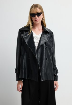 Vrouw in een oversized zwarte faux leren jack, witte blouse, zwarte wijde broek, kattenogen zonnebril en grote hoop oorbellen tegen een witte achtergrond.