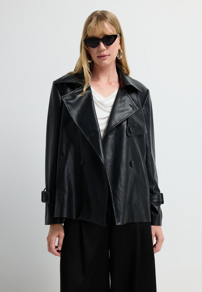 Donna con giacca oversize in ecopelle nera, blusa bianca, pantaloni larghi neri, occhiali cat-eye e grandi orecchini a cerchio, su sfondo bianco.