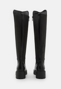 Bottes noires à talons hauts, montant jusqu'au genou, avec une finition en cuir lisse et des panneaux en tissu texturé, dotées de zips latéraux et d'une semelle épaisse et robuste.
