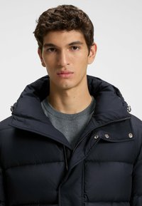 Marinblå puffjacka med hög krage, knappdetaljer och dragkedjestängning, bärs över en grå sweatshirt med rund hals. Slät, isolerad tyg.