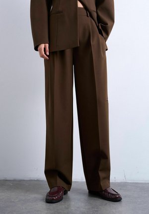 Person iført brune højttaljede bredbenede bukser, matchende brun blazer og mørkebrune loafers, står på en beton gulv.