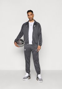 Mand i grå Chicago Bulls træningsdragt, der holder en sort og grå Jordan basketball, stående mod en helt hvid baggrund.