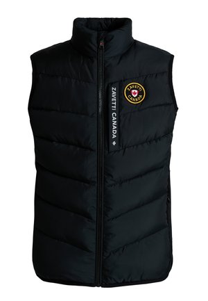 Kind mit geflochtenem Haar, das eine schwarze Steppweste, schwarzen Pullover, schwarze Hose und schwarze Sneakers trägt und vor einem grauen Hintergrund steht.