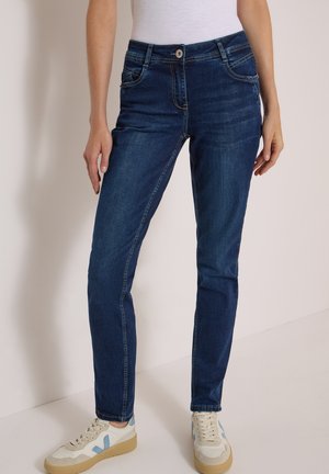 Personne portant un jean slim bleu foncé et des baskets blanches avec des touches bleues et beiges, debout devant un fond clair uni.