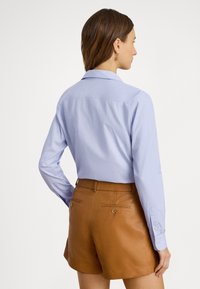 Lyseblå skjorte med lange ermer og spiss krage, med mansjetter med knapper, kombinert med brune skinnshorts med baklommer.