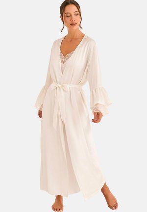 Femme portant une longue robe blanche avec des poignets en dentelle et une ceinture nouée à la taille, debout pieds nus devant un fond uni.