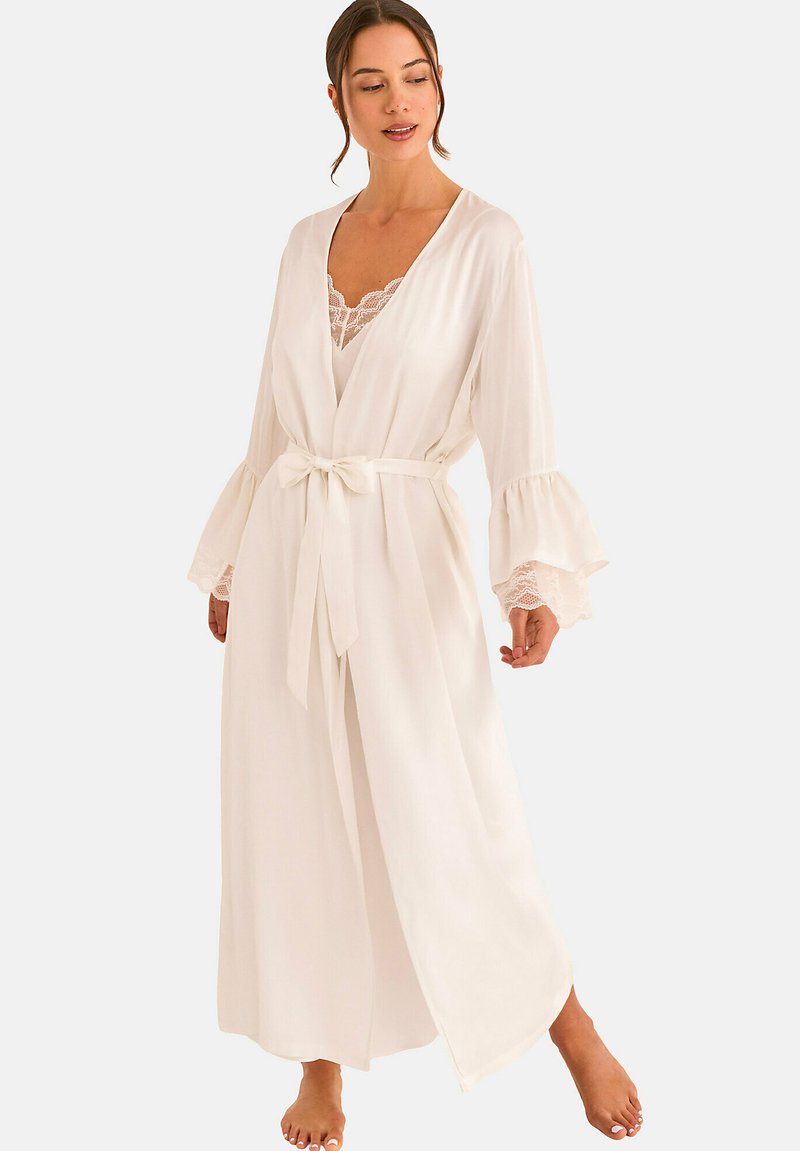 Femme portant une longue robe blanche avec des poignets en dentelle et une ceinture nouée à la taille, debout pieds nus devant un fond uni.