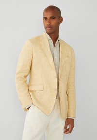 Blazer de lino en amarillo claro con un ajuste entallado, cierre de un botón, solapas de muesca y bolsillos laterales. Llevado sobre una camisa beige.