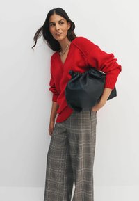 Red knitted sweater with a V-neck, paired with gray plaid wide-leg trousers. Model holds a black leather drawstring bag. Plain background.