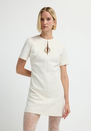 Donna con capelli biondi corti, che indossa un vestito avorio a maniche corte con una spilla dorata e collant bianchi a fantasia, in piedi su uno sfondo bianco semplice.