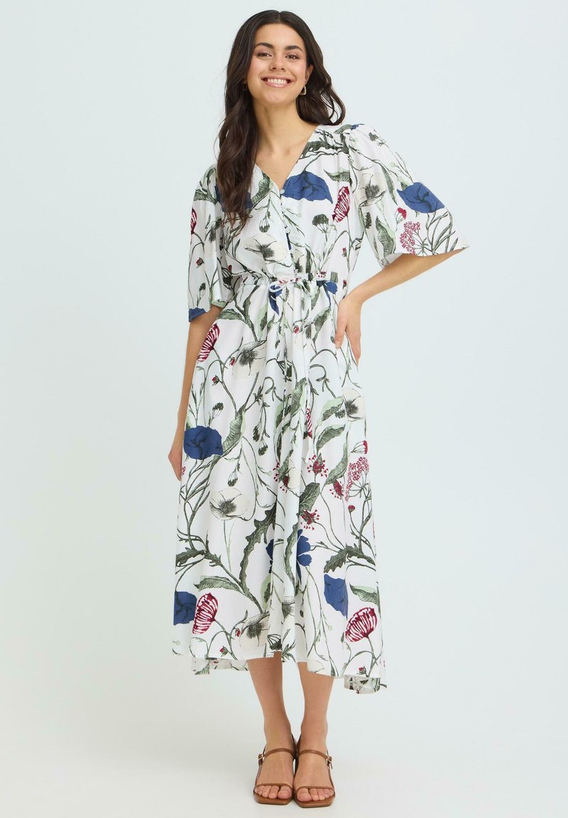 Femme souriante portant une robe midi blanche avec des motifs floraux bleus, rouges et verts, posant avec une main sur la hanche et portant des sandales marron.